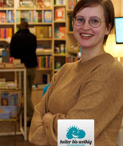 Frau mit Brille und braunem Pullover lächelt in einer Buchhandlung, im Hintergrund Bücherregale.
