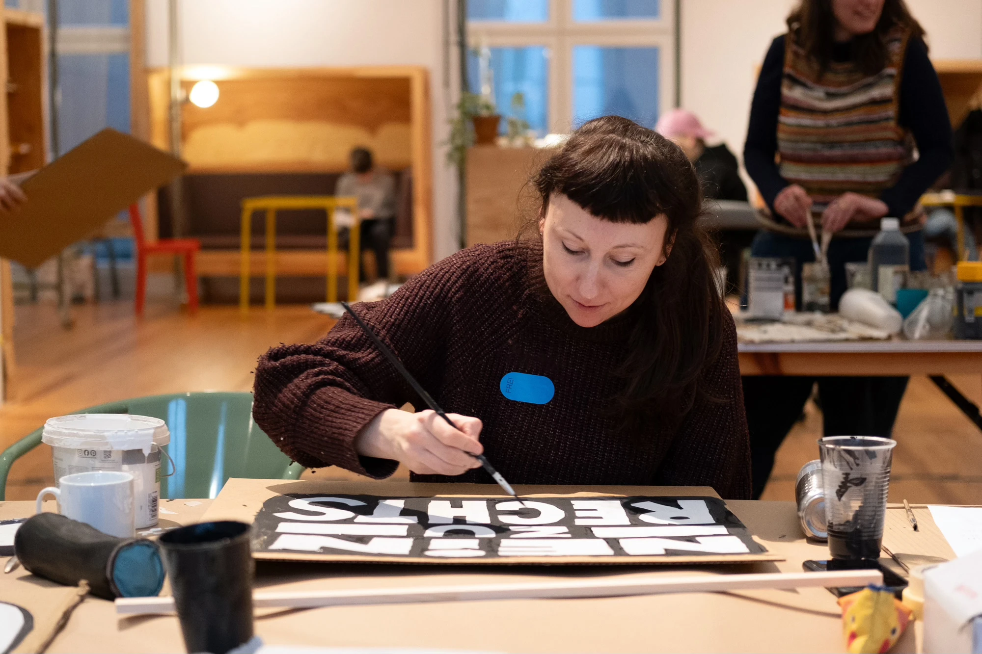 Eine Person mit langen Haaren sitzt an einem Tisch und malt ein Demoschild.